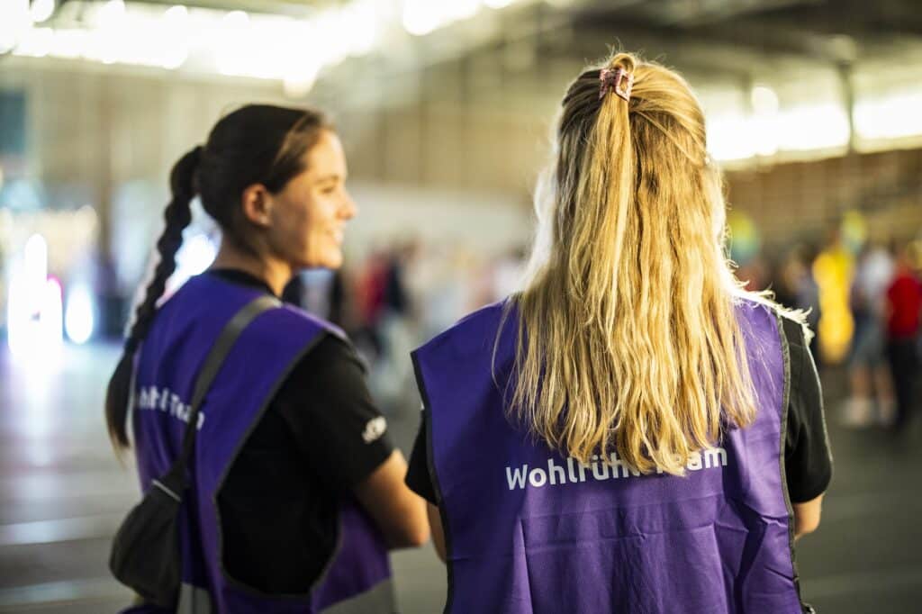 Zwei Mitglieder des „Wohlfühlteams“ in lila Westen stehen vor einer Veranstaltung.